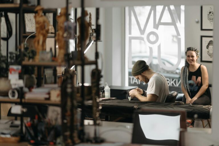 Tattoo artist creating design in a modern studio with client waiting. Professional workspace and tools visible.